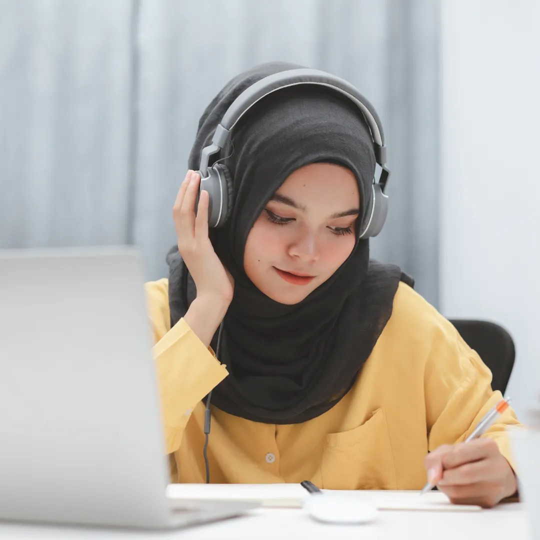 Person wearing headphones sits at a desk, writing notes, with a laptop.