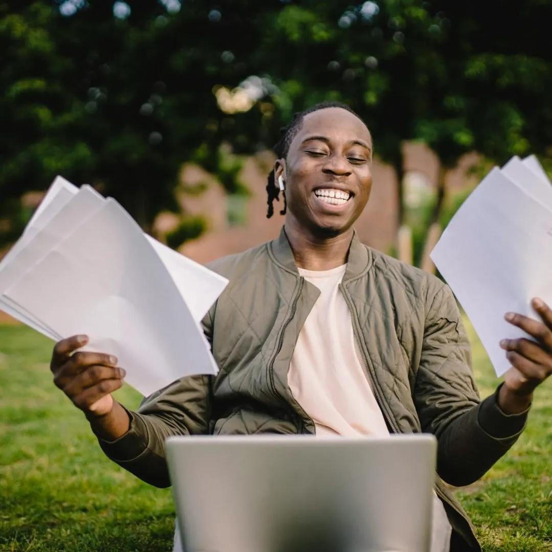 A person sits on grass, holding multiple sheets of paper in each hand.